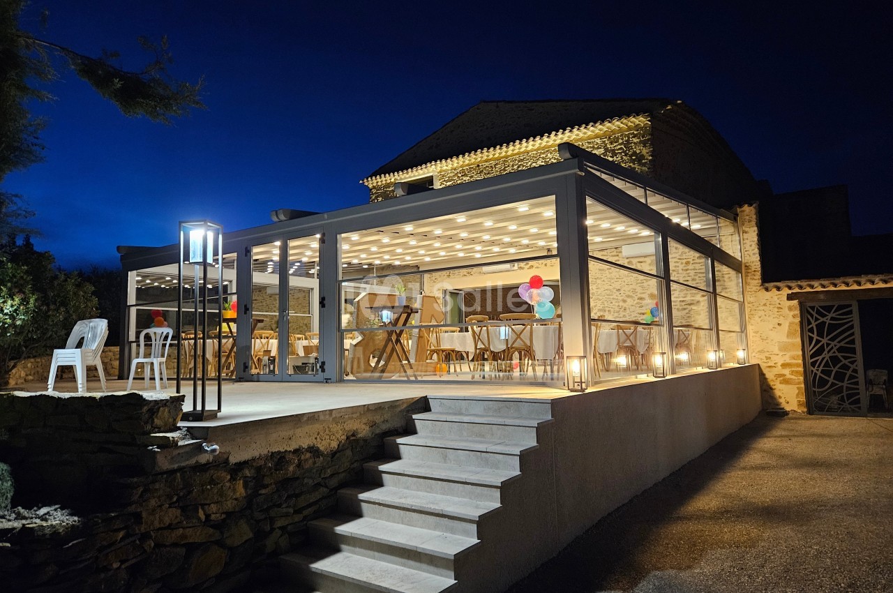 Terrasse éclairée avec une structure vitrée, décorée pour un événement en soirée, devant un bâtiment en pierre.