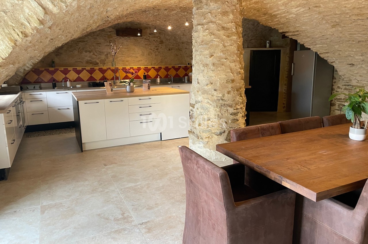 Cuisine voûtée en pierre avec meubles modernes, table en bois, chaises marron et crédence colorée.