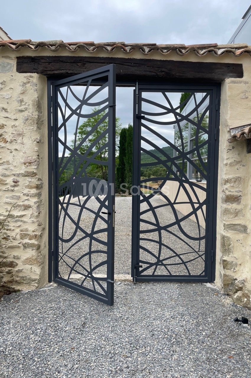 Portail métallique ajouré au design moderne, installé entre deux murs en pierre, donnant sur une allée gravillonnée.
