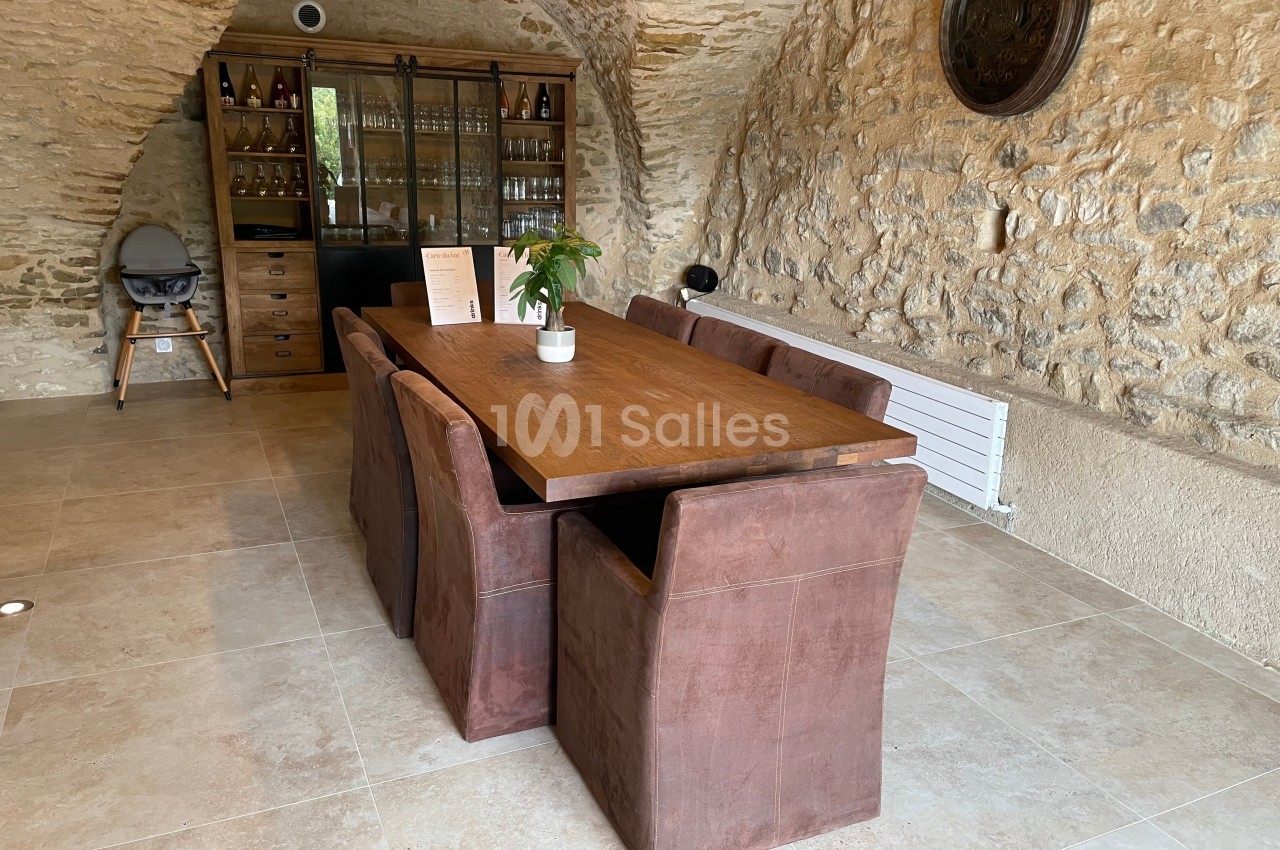 Table en bois entourée de chaises marron dans une pièce en pierre avec un buffet vitré en arrière-plan.