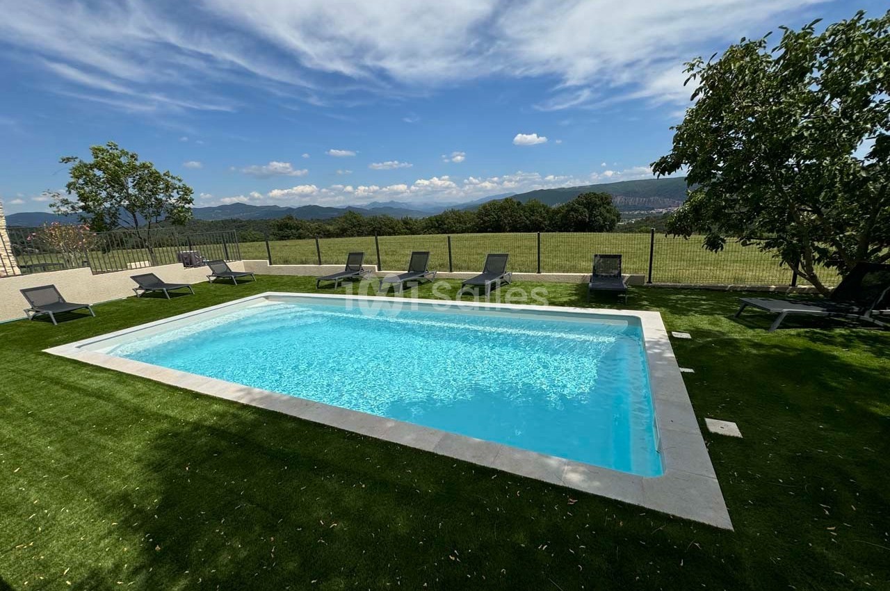 Piscine rectangulaire entourée de chaises longues sur pelouse artificielle, avec vue sur un paysage vallonné.