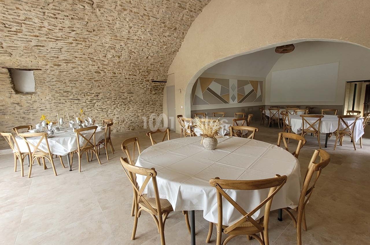 Salle voûtée en pierre avec tables rondes dressées de nappes blanches et chaises en bois, ambiance lumineuse et épurée.