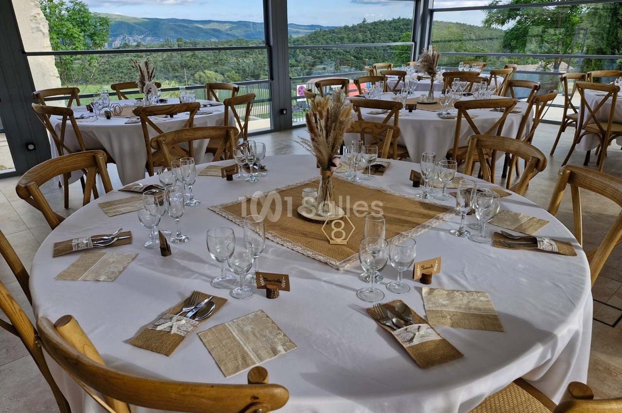 Tables rondes dressées avec nappes blanches, décor champêtre et vue sur un paysage vallonné à travers de grandes baies…