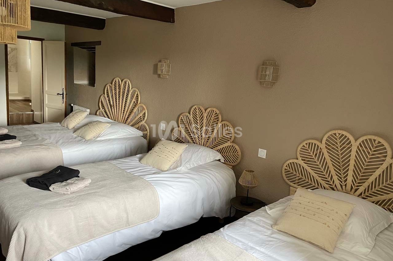 Chambre avec trois lits simples, têtes de lit en rotin, murs beige, poutres apparentes et lumière naturelle.