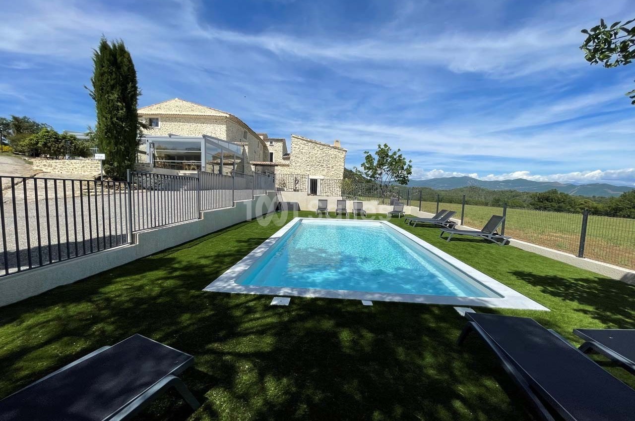 Piscine extérieure entourée de chaises longues sur une pelouse, avec vue sur une maison en pierre et des montagnes.