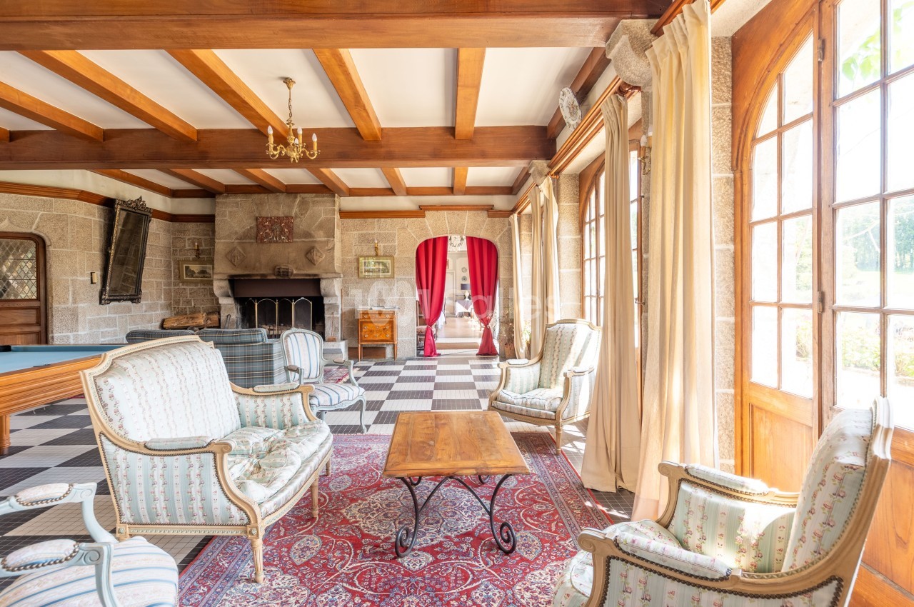 Salon lumineux avec poutres apparentes, cheminée en pierre, mobilier classique et grandes fenêtres donnant sur l'extérieur.