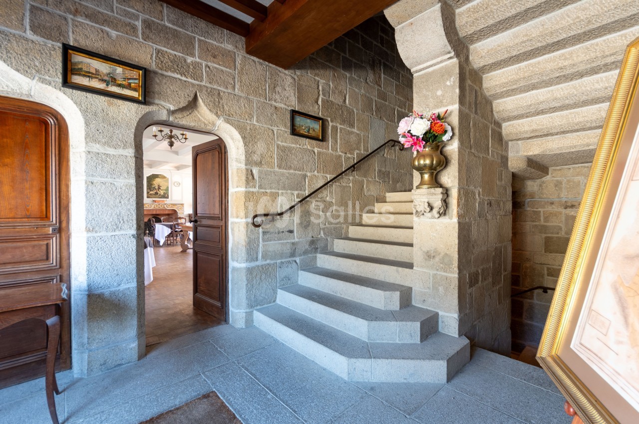 Escalier en pierre dans un intérieur ancien, avec une rampe en métal et un vase de fleurs sur une colonne.
