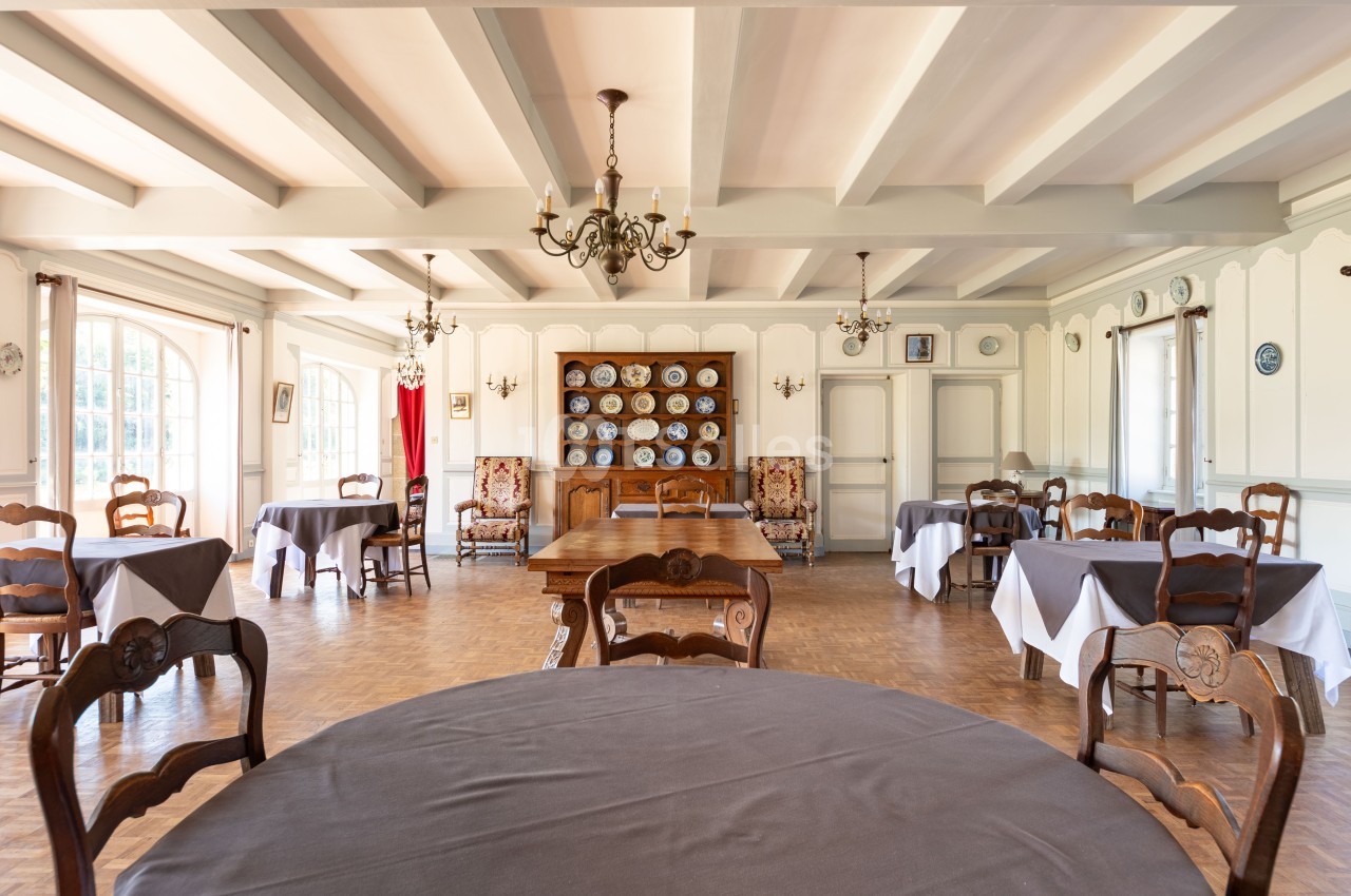 Salle à manger lumineuse avec tables recouvertes de nappes grises, chaises en bois et vaisselier en arrière-plan.
