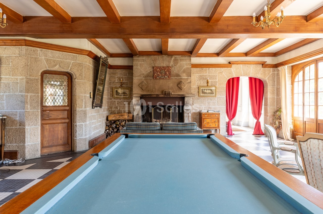 Salle avec table de billard, cheminée en pierre, poutres apparentes et grandes fenêtres avec rideaux rouges.