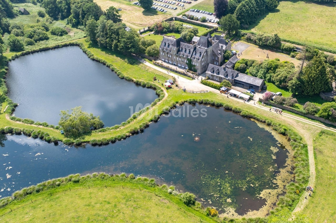 Vue aérienne d'un manoir entouré de deux étangs et de verdure, avec un parking visible en arrière-plan.