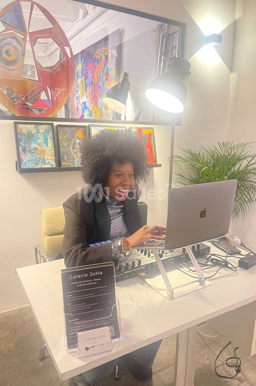 Femme souriante travaillant sur un ordinateur portable dans une galerie d'art, entourée de peintures et d'une plante.