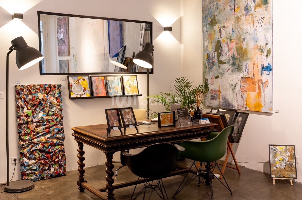 Espace d'exposition artistique avec une table en bois, des chaises, des tableaux colorés et un grand miroir mural.
