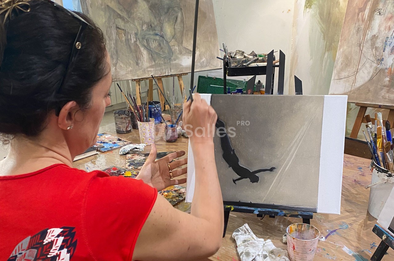 Une femme peint une silhouette sombre sur une toile dans un atelier artistique, entourée de pinceaux et de peintures.