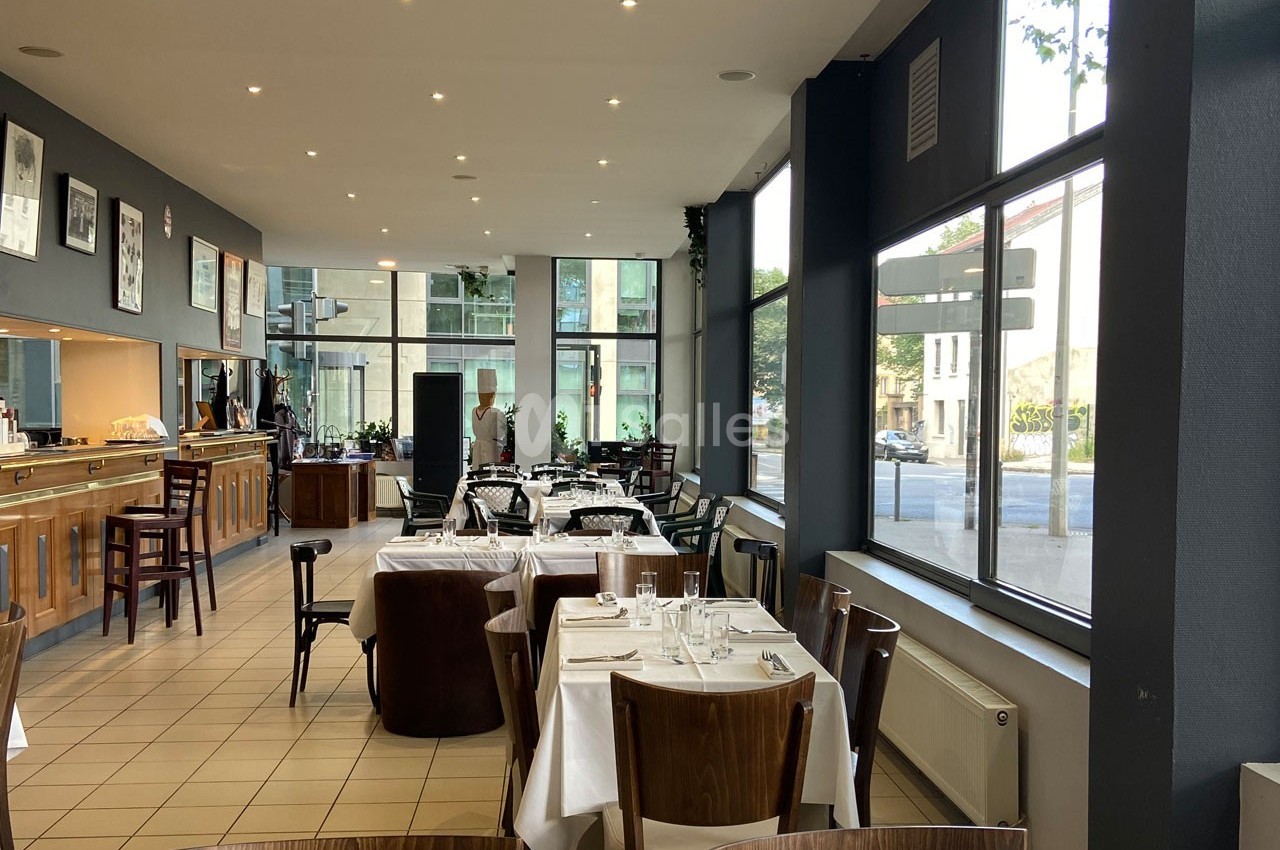 Salle de restaurant lumineuse avec des tables dressées, grandes baies vitrées et vue sur la rue.