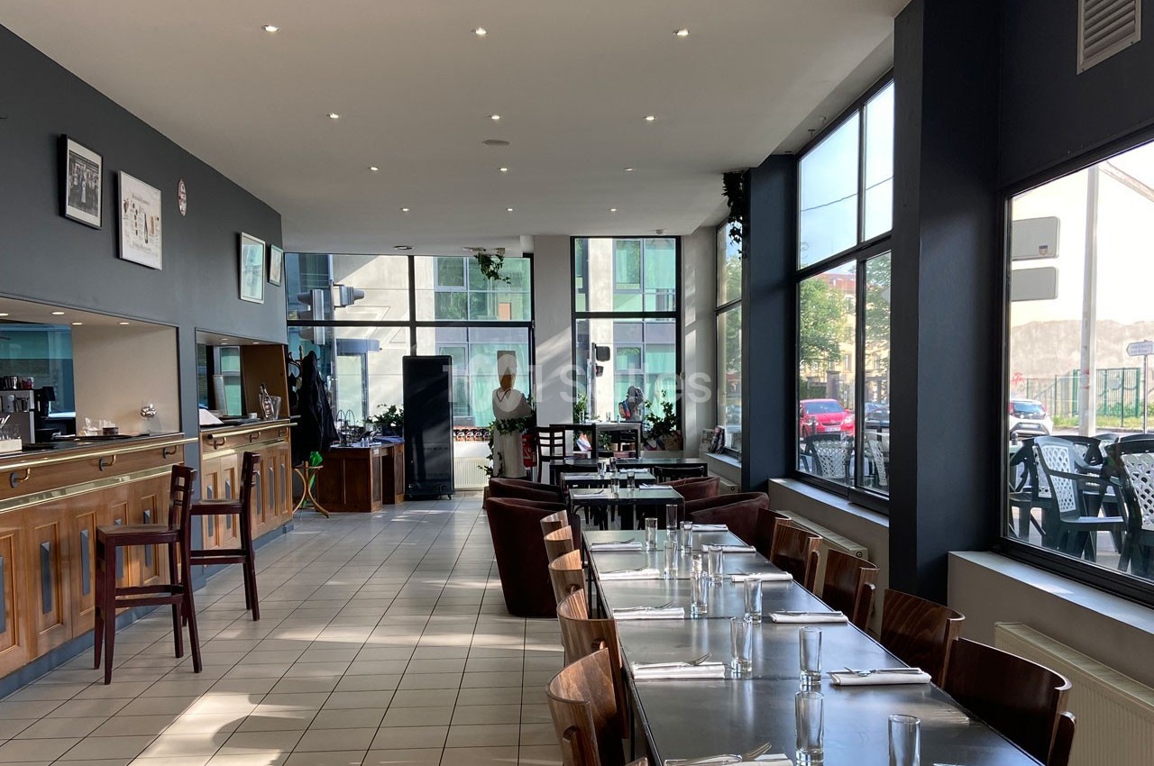 Salle de restaurant lumineuse avec grandes baies vitrées, tables dressées et comptoir en bois à gauche.