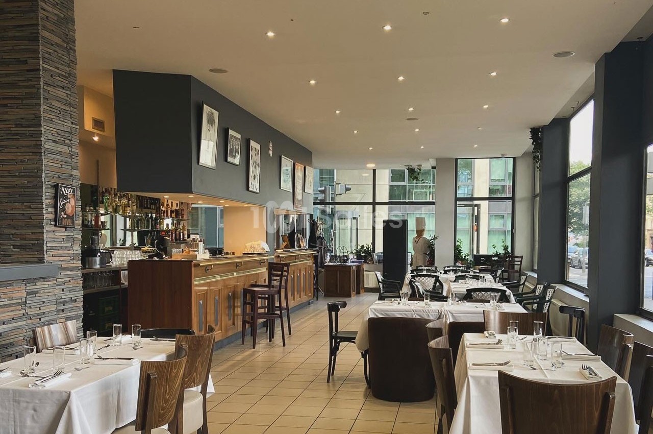Salle de restaurant lumineuse avec tables dressées, bar en bois et grandes baies vitrées donnant sur l'extérieur.