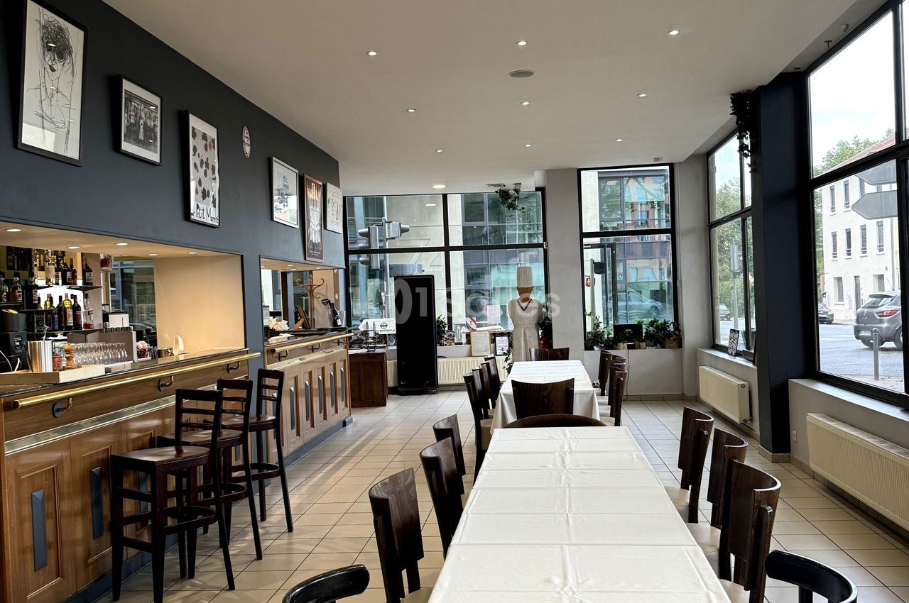 Salle de restaurant lumineuse avec tables alignées, chaises en bois, bar à gauche et grandes fenêtres donnant sur l…