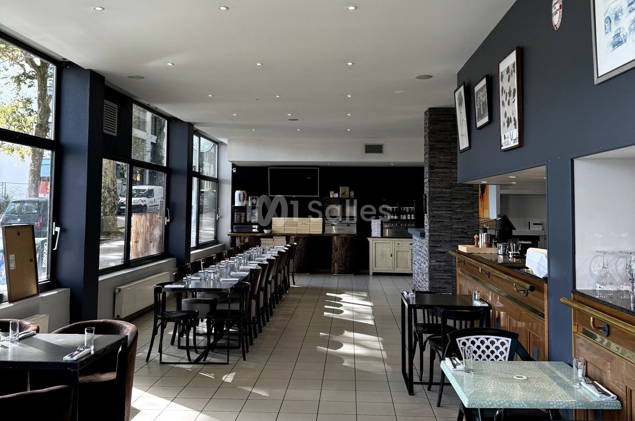 Salle de restaurant lumineuse avec tables dressées, grandes baies vitrées et comptoir en bois à droite.
