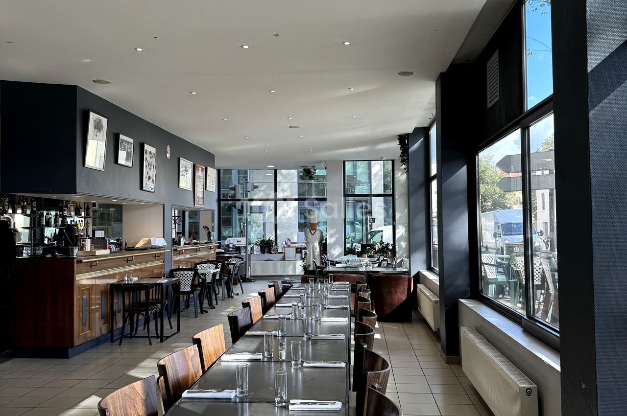 Salle de restaurant lumineuse avec tables dressées, grandes baies vitrées et un comptoir en bois au fond.