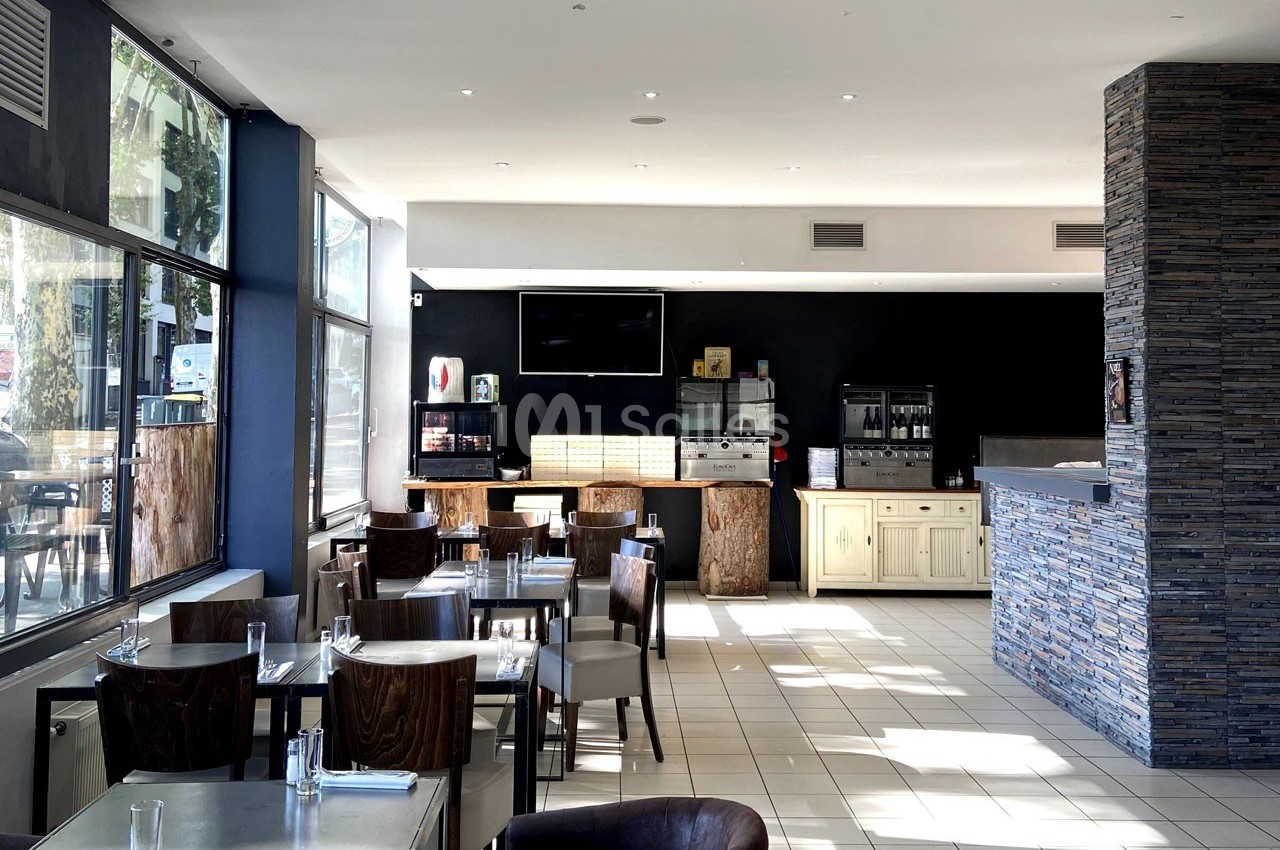 Salle de restaurant lumineuse avec tables dressées, grandes baies vitrées et comptoir en pierre au fond.