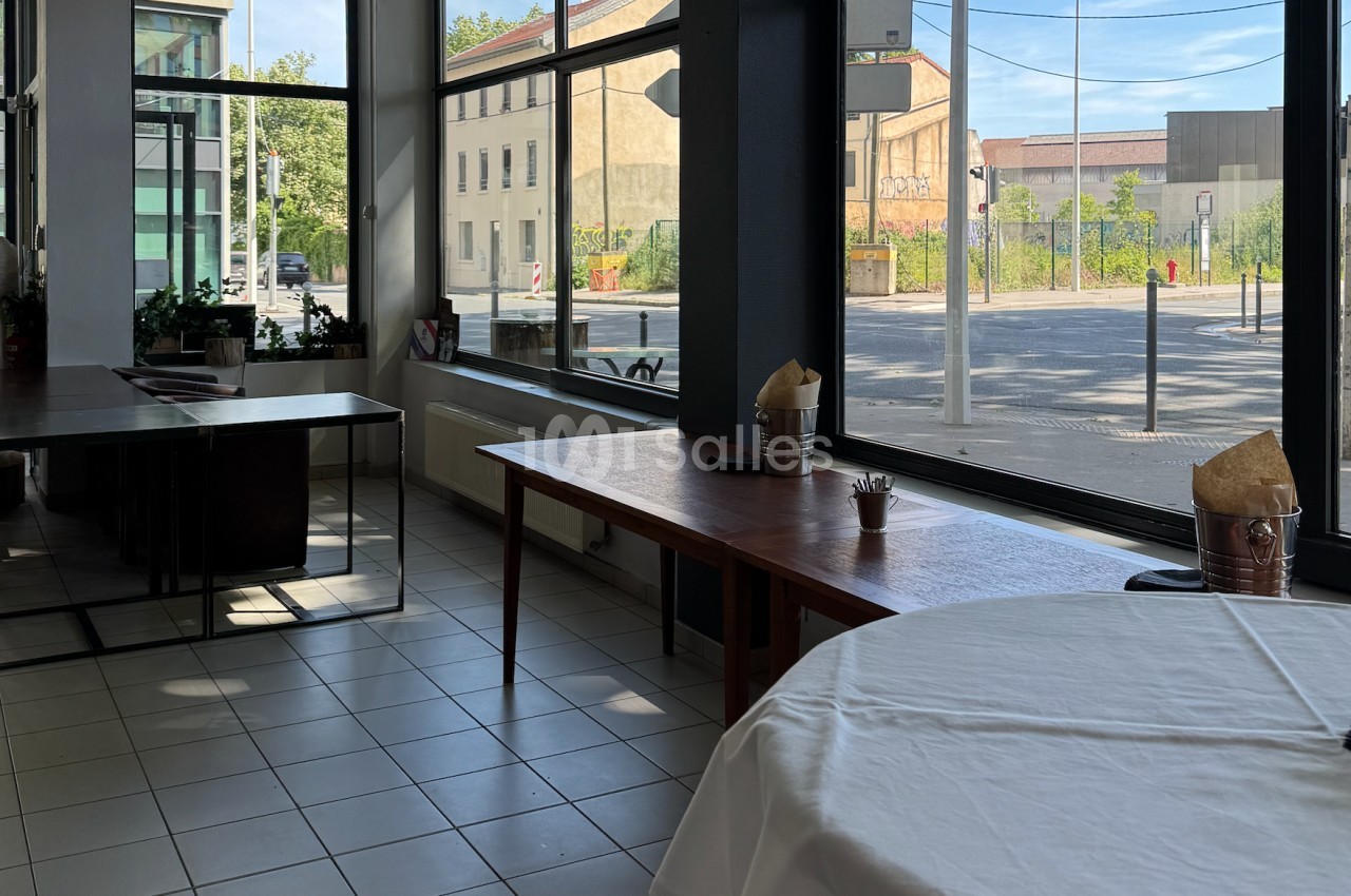 Salle lumineuse avec grandes baies vitrées, tables en bois et vue sur une rue calme bordée de bâtiments.