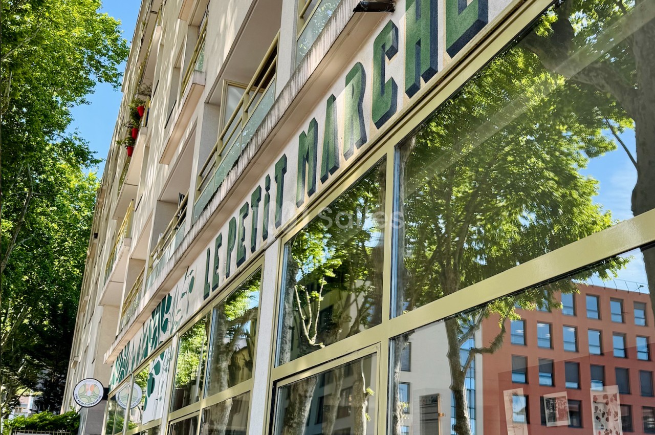Façade d'un immeuble avec une devanture vitrée et l'inscription ’Marché’ reflétant des arbres et des bâtiments voisins.