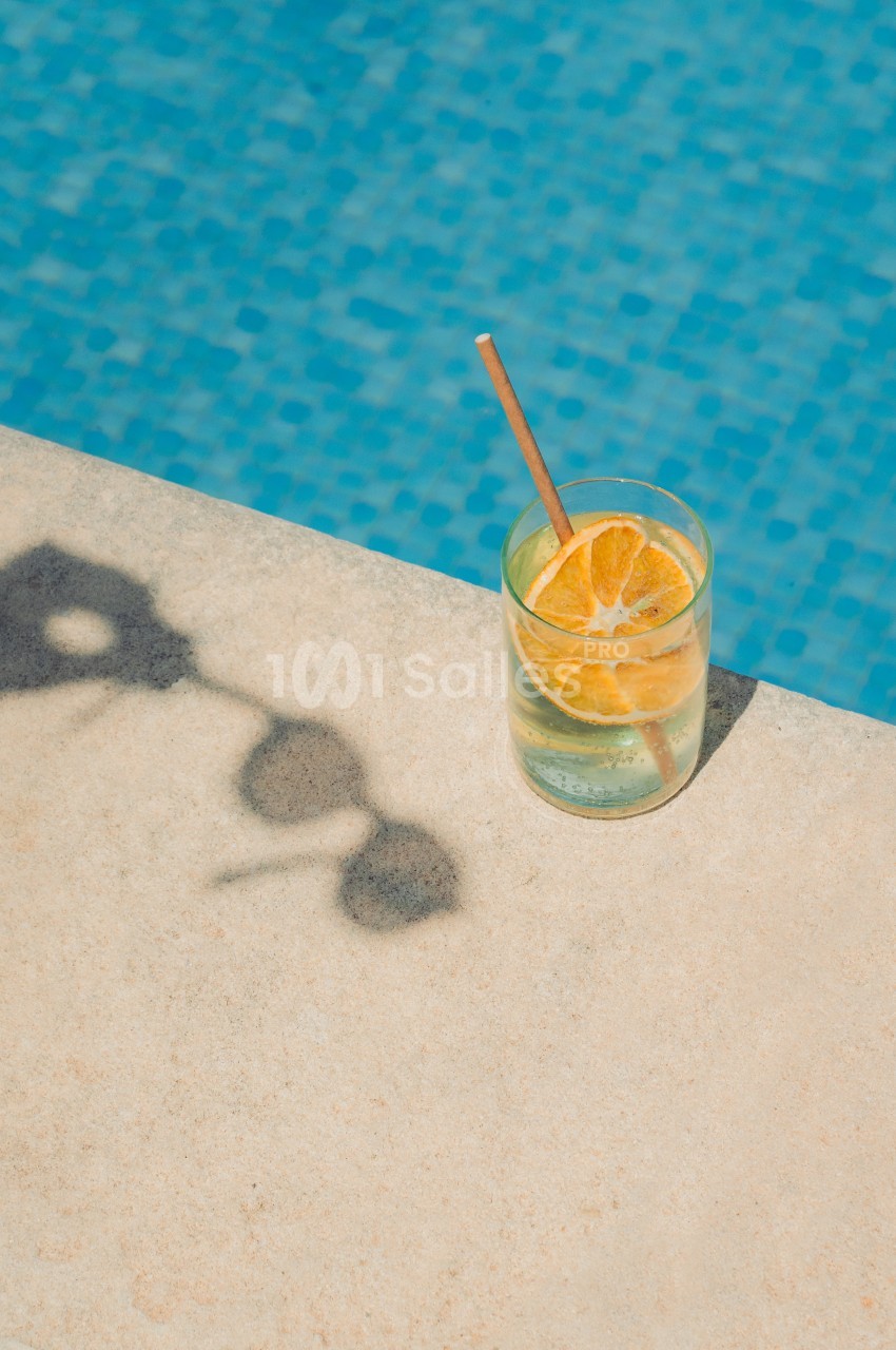 Verre de boisson fraîche avec des tranches d'orange et une paille, posé sur le bord d'une piscine en mosaïque bleue.