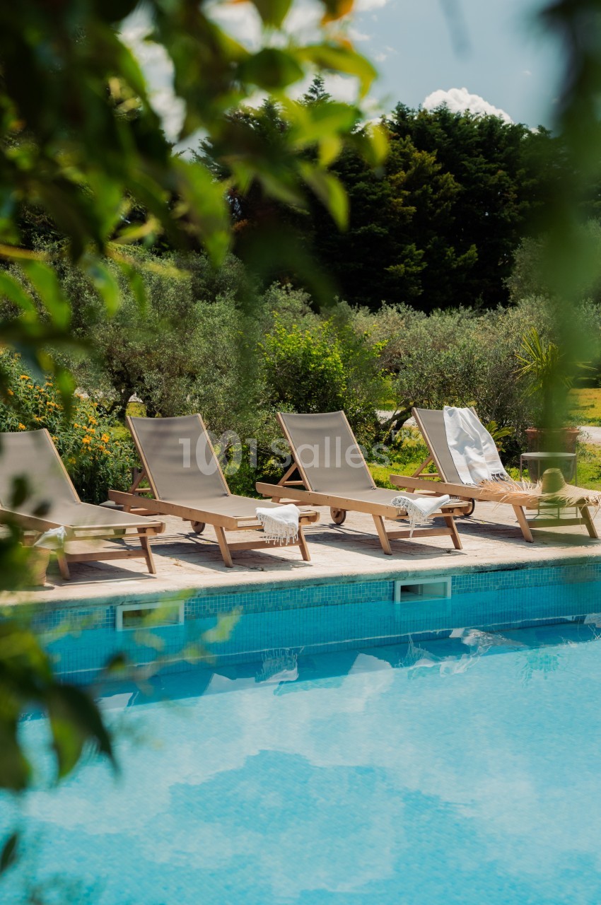 Chaises longues alignées au bord d'une piscine, entourées de végétation et d'arbres sous un ciel dégagé.