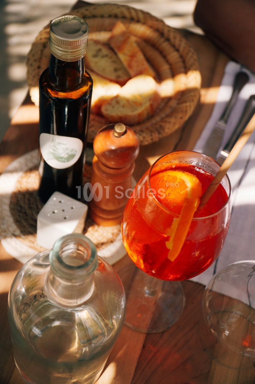 Verre de cocktail orange avec une tranche d'orange, entouré de pain, huile, poivrier et carafe sur une table en bois.