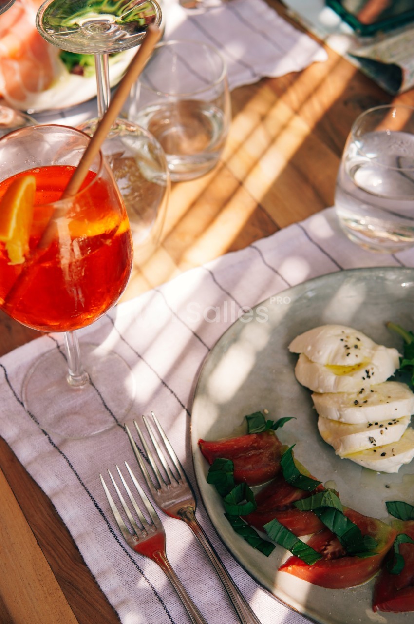 Assiette avec mozzarella, tomates et basilic, accompagnée d'un verre de cocktail orange sur une table ensoleillée.