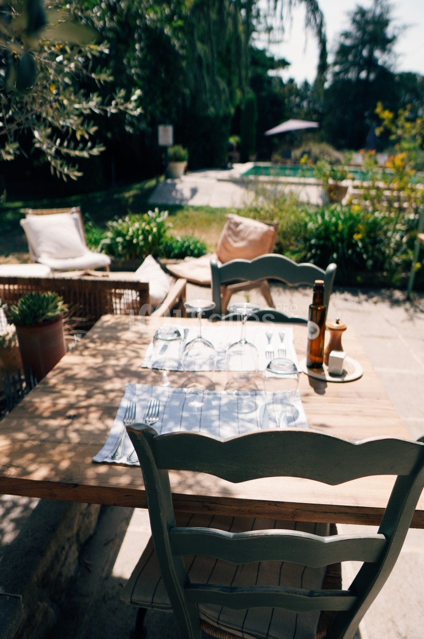 Table en bois dressée avec des couverts et des verres, située en extérieur près d'un jardin et d'une piscine.