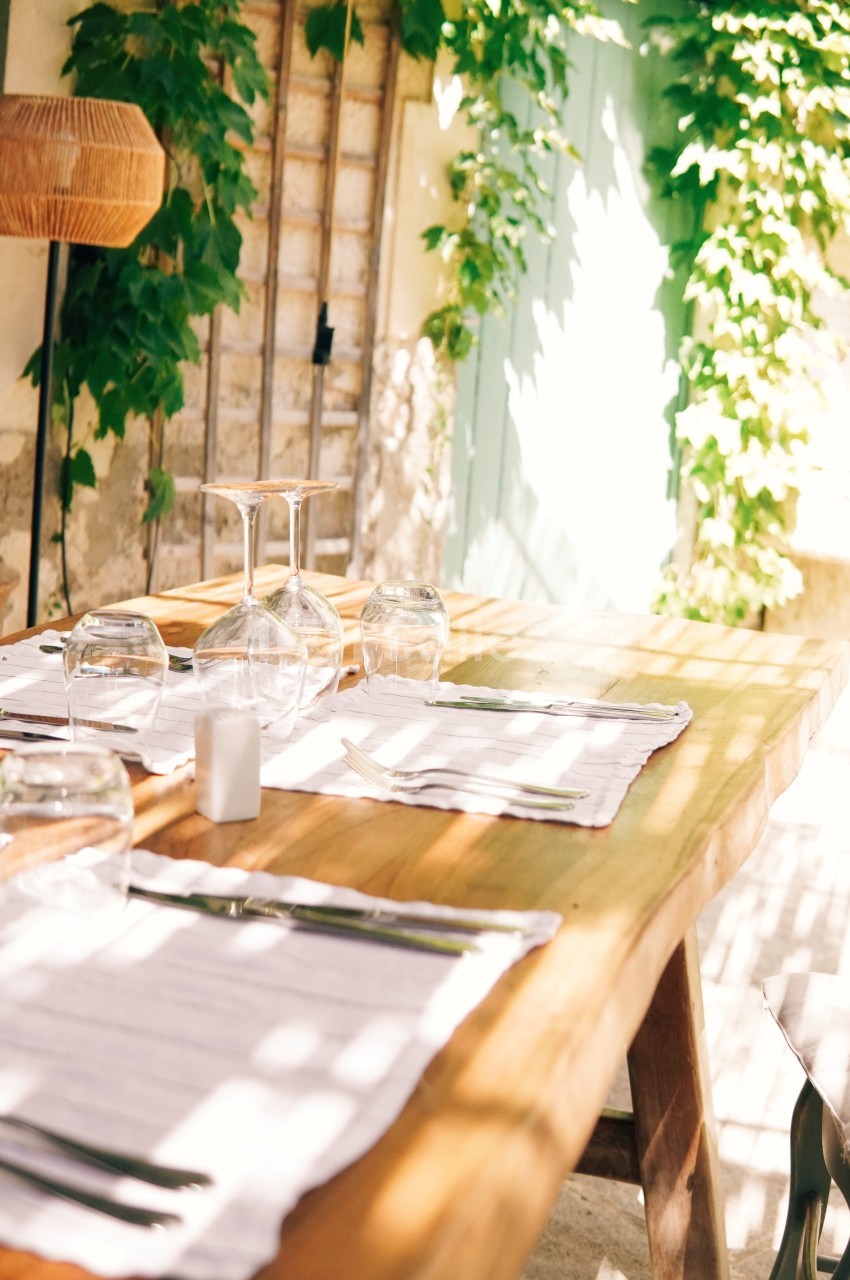 Table en bois dressée avec des verres et des couverts, située en extérieur près d'un mur couvert de plantes grimpantes.