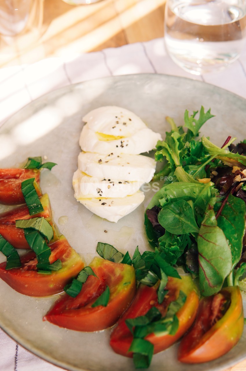 Assiette avec tranches de tomates, mozzarella assaisonnée et salade verte sur une table lumineuse.