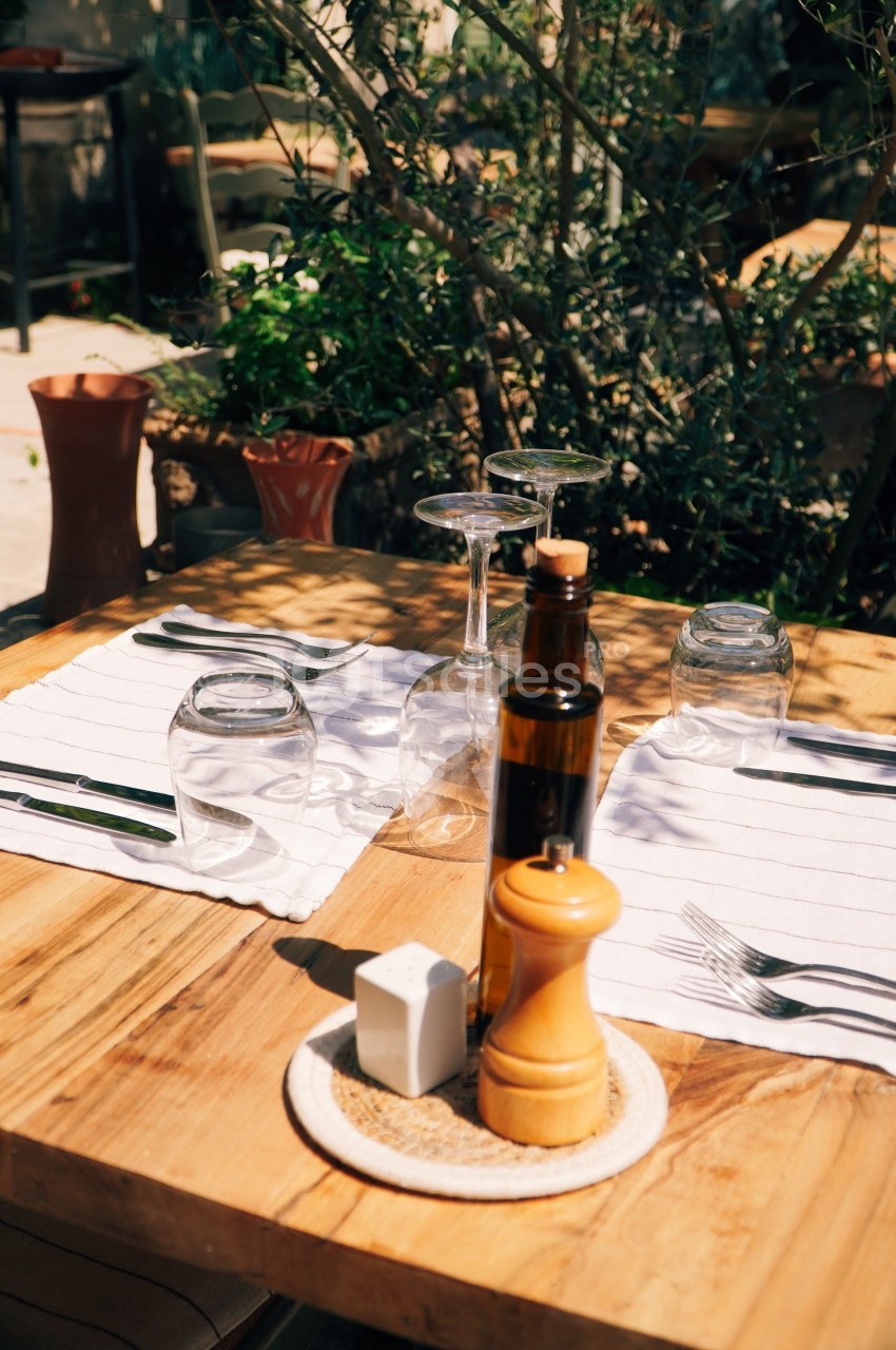 Table en bois dressée avec couverts, verres, huile, vinaigre, sel et poivre, entourée de plantes en extérieur.