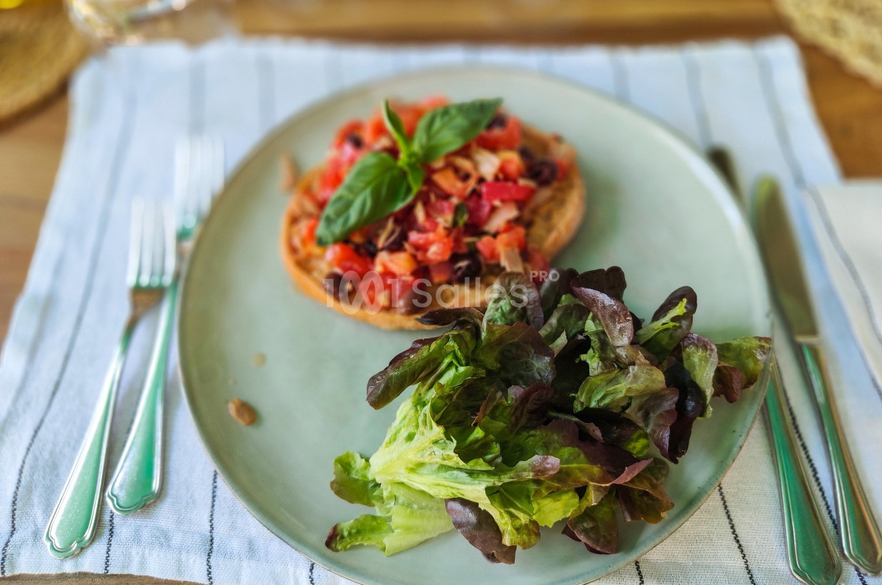 Tartine garnie de tomates, olives et basilic, accompagnée de salade verte sur une assiette posée sur une nappe rayée.
