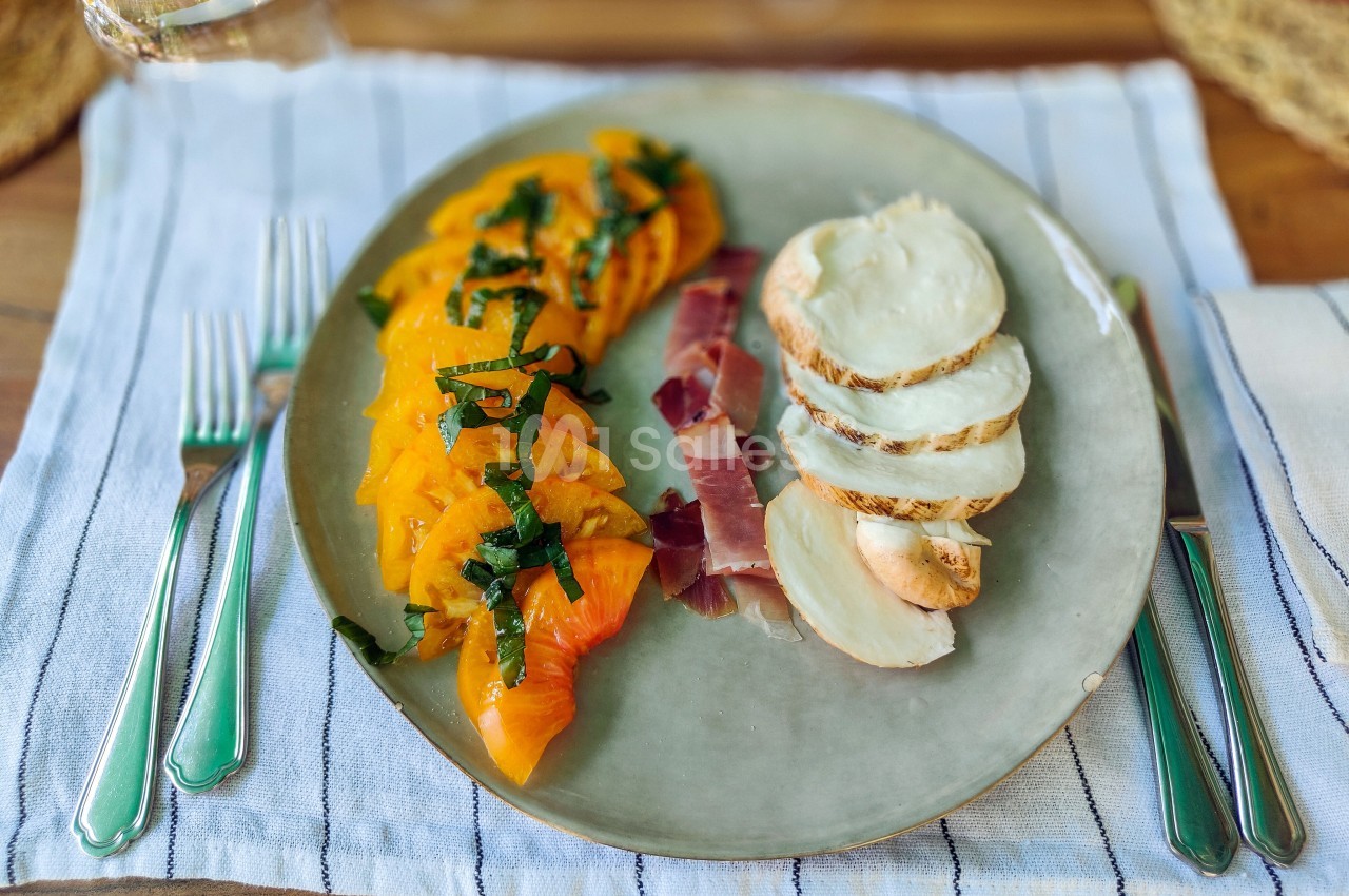 Assiette contenant des tranches de tomates jaunes, mozzarella, jambon cru et basilic, sur une nappe rayée.