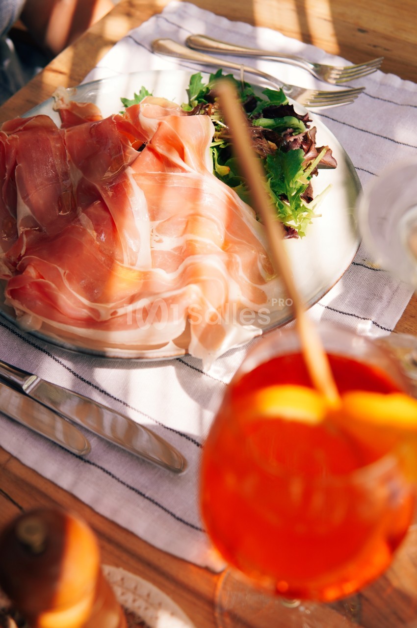 Assiette de jambon cru accompagnée de salade verte, servie sur une nappe rayée avec un verre de cocktail orange.