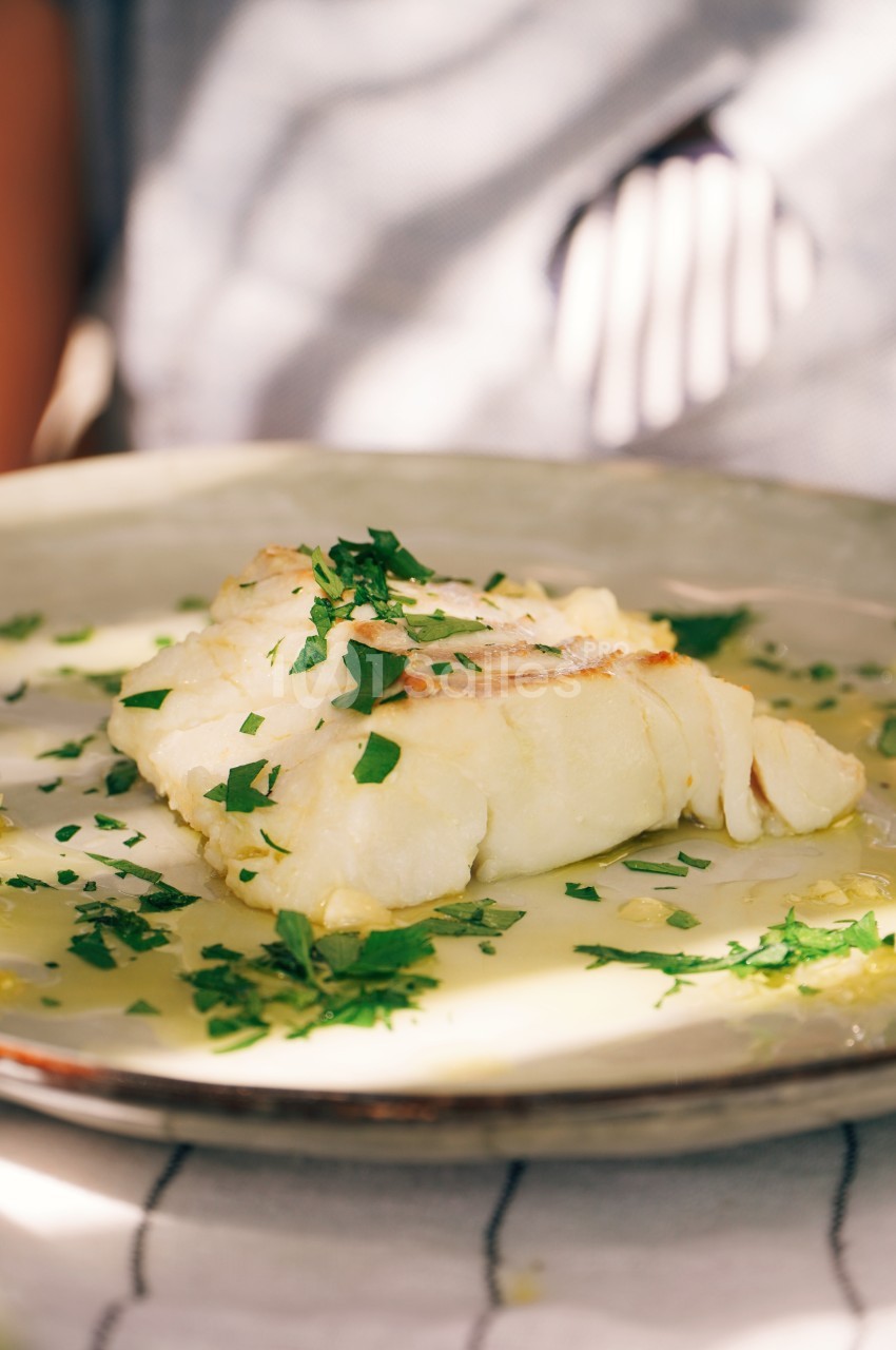 Filet de poisson blanc servi avec une sauce au beurre et parsemé de persil frais sur une assiette en céramique.