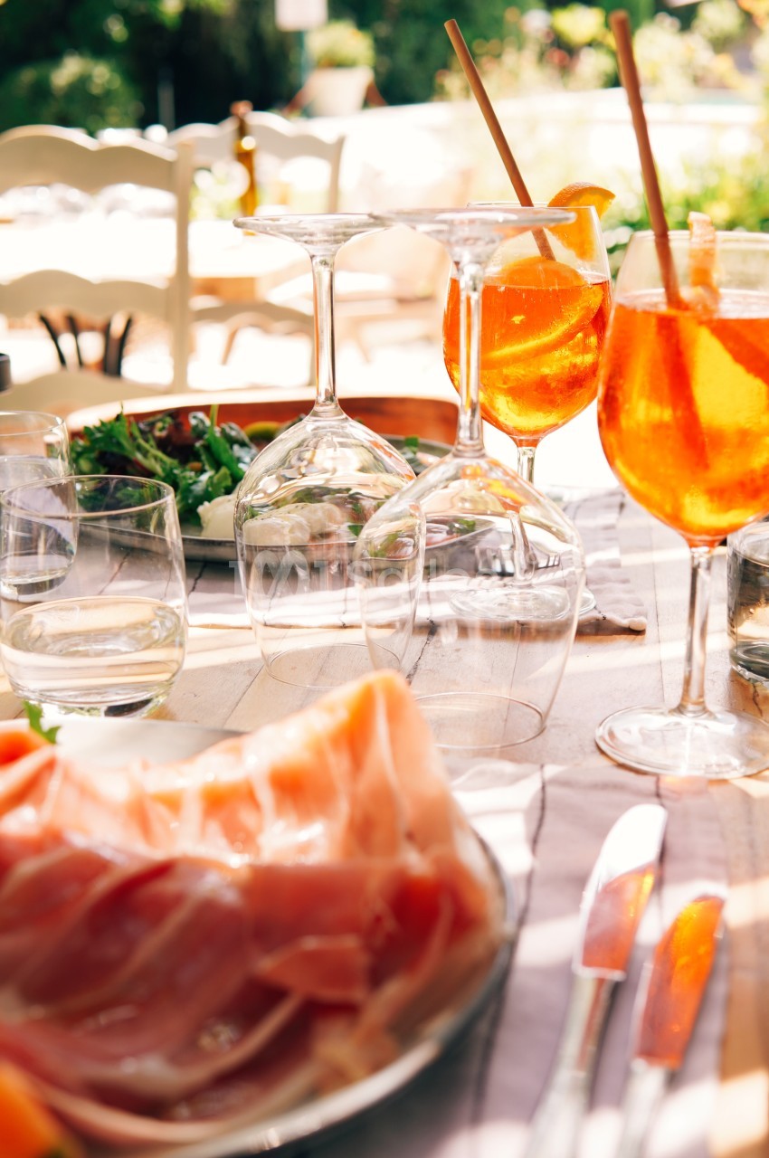Table dressée en extérieur avec des verres de spritz, une assiette de jambon cru et une salade verte.