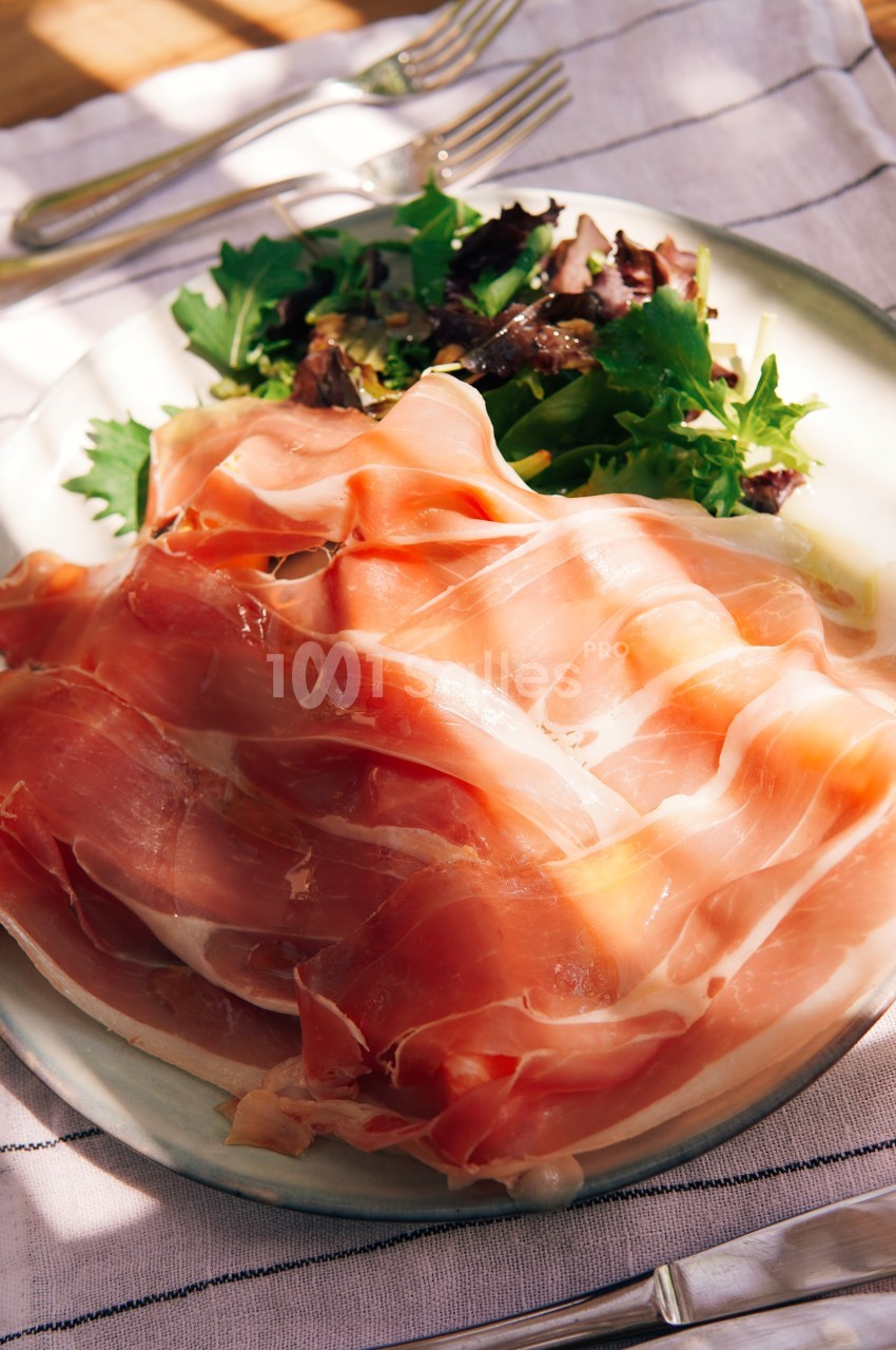 Assiette de tranches de jambon cru accompagnées d'une salade verte sur une nappe blanche.