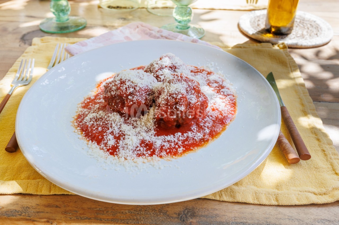 Assiette de boulettes de viande nappées de sauce tomate et saupoudrées de fromage râpé, posée sur une table ensoleillée.