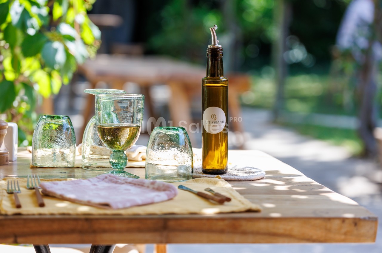 Bouteille d'huile d'olive, verres et serviette disposés sur une table en bois dans un cadre extérieur verdoyant.
