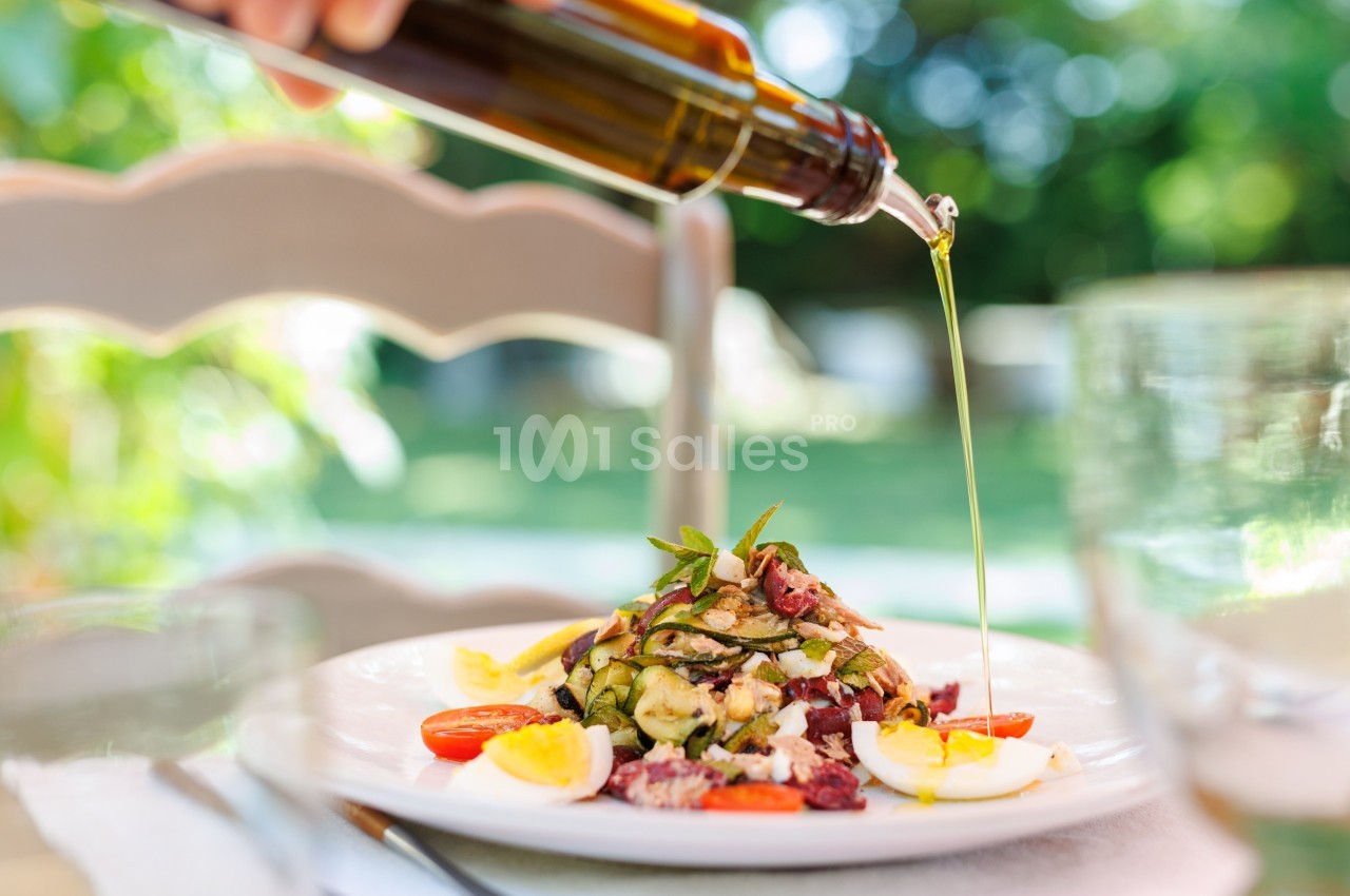 Assiette de salade garnie de légumes, œufs et tomates, arrosée d'huile d'olive versée depuis une bouteille.