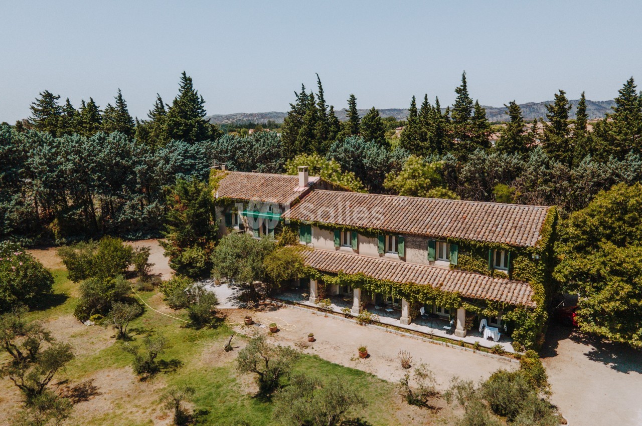 Vue aérienne d'une maison en pierre entourée d'arbres et d'un jardin, sous un ciel clair.