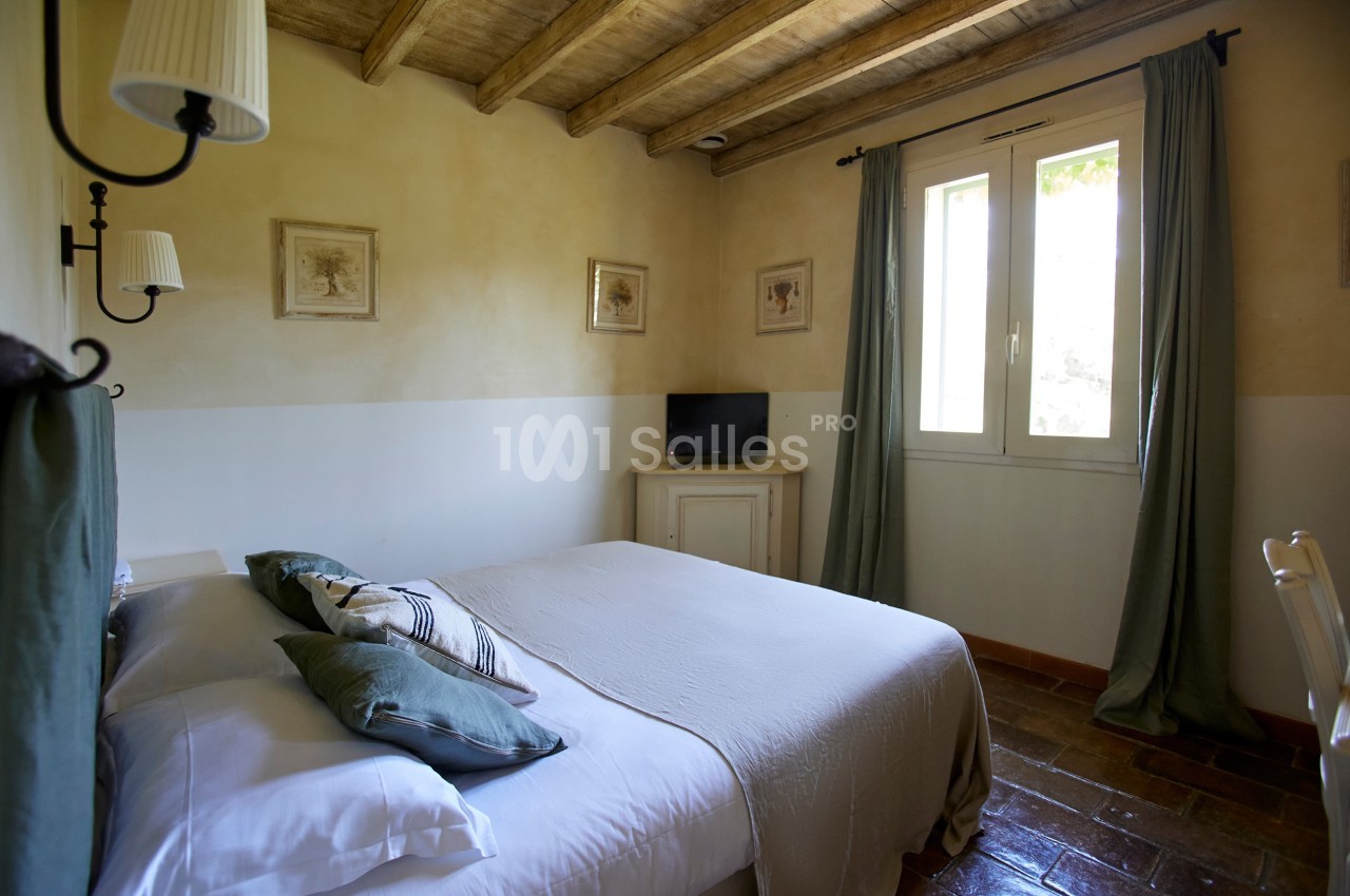 Chambre lumineuse avec lit double, draps blancs, coussins verts, télévision, fenêtres avec rideaux et plafond en bois.