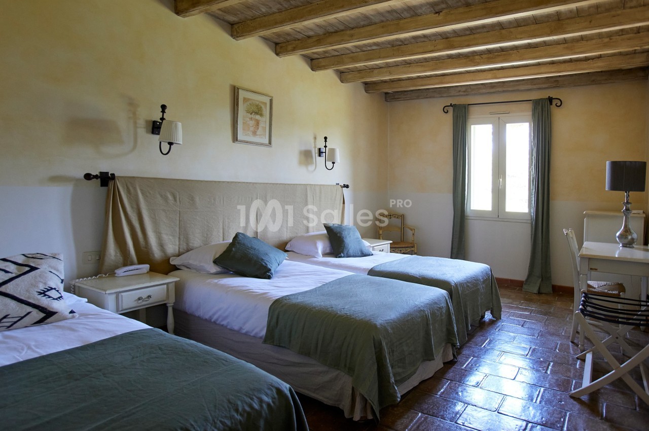 Chambre lumineuse avec trois lits simples, murs clairs, plafond en poutres apparentes et fenêtre avec rideaux verts.