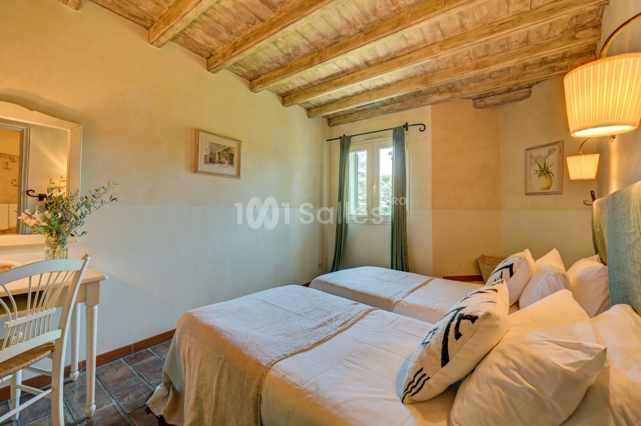 Chambre lumineuse avec deux lits simples, plafond en bois, murs clairs, bureau et fenêtre donnant sur un extérieur verdoyant.
