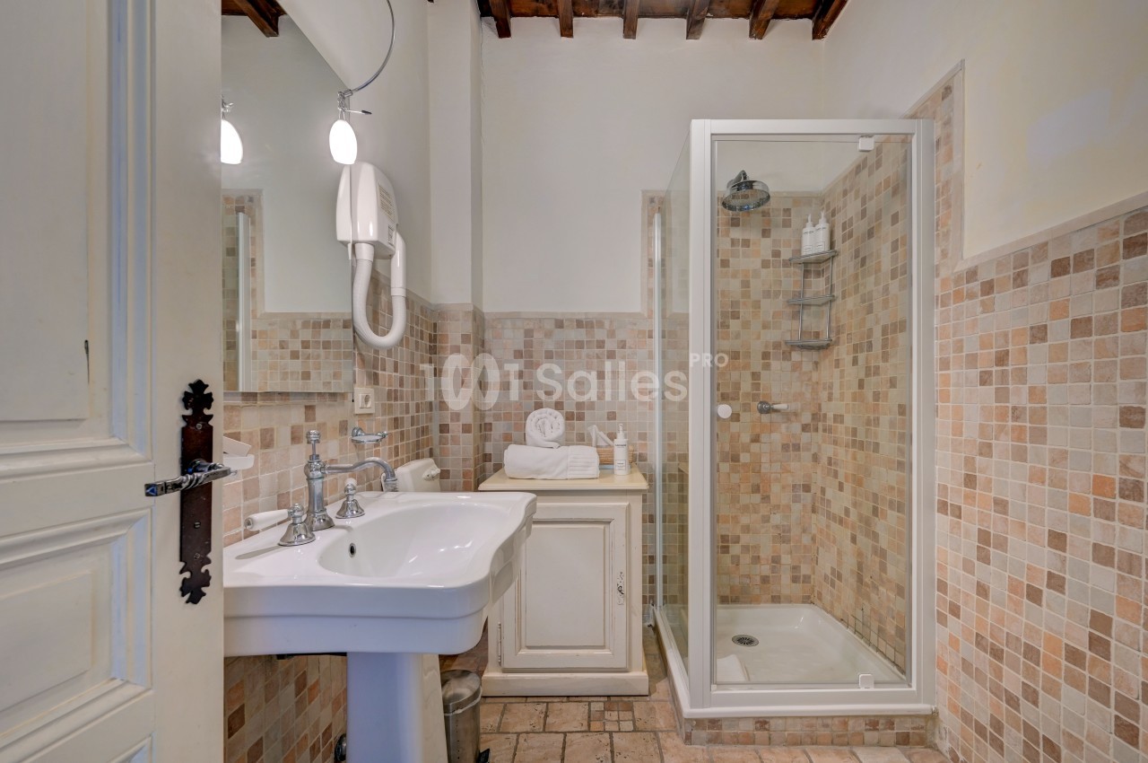 Salle de bain avec douche à l'italienne, lavabo blanc, carrelage beige et miroir sous un plafond en bois.