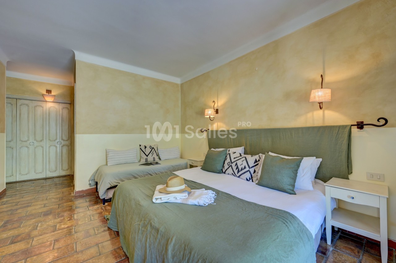Chambre lumineuse avec un lit double, un lit simple, des murs beige clair, et un sol en carrelage.