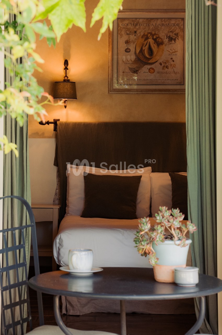 Vue d'une chambre cosy avec lit, éclairage doux, rideaux verts et table extérieure ornée de plantes et d'une tasse.